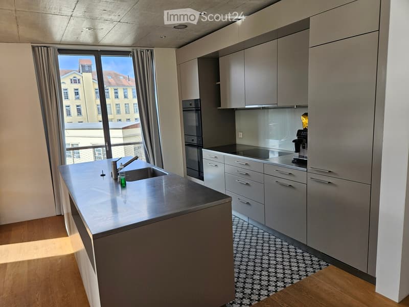 Modern kitchen with grey cabinets, stainless steel sink, oven, coffee machine, and large window with view.