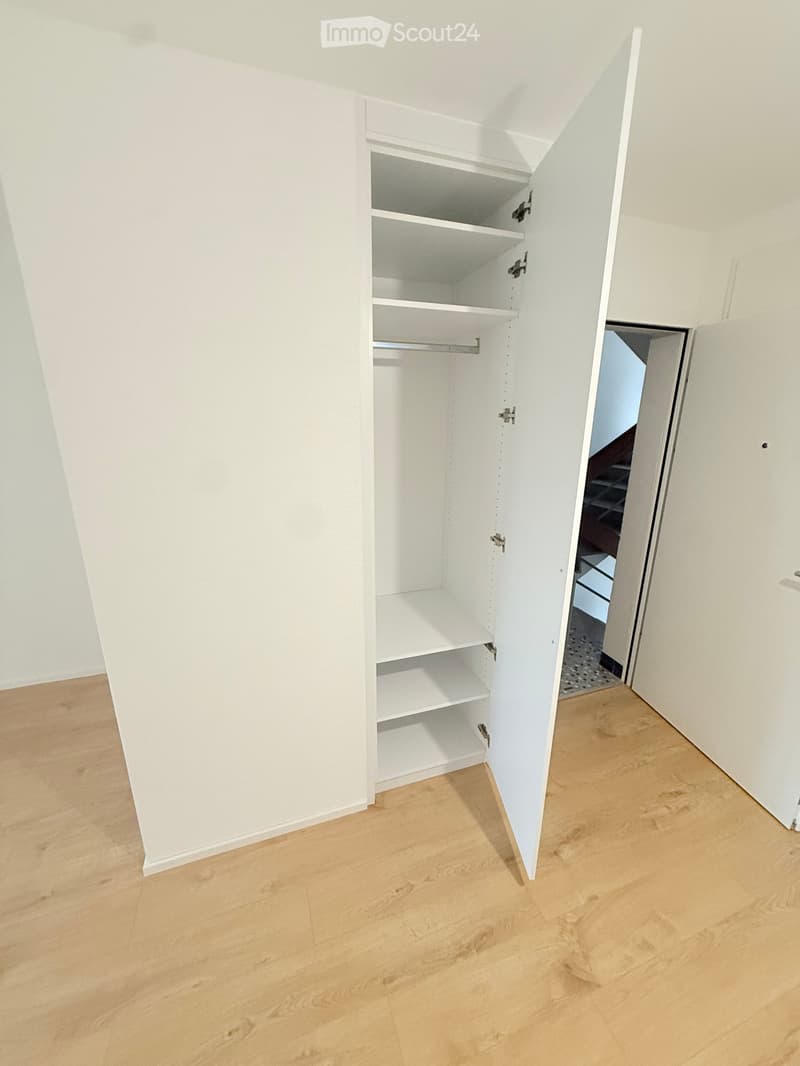 Empty room with light wooden flooring, white walls, and an open closet with empty shelves and a hanging rod.