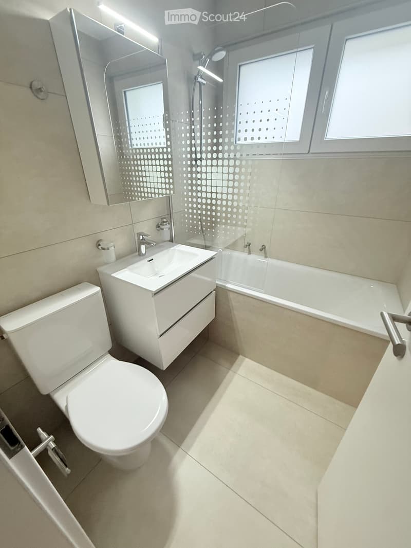 Modern bathroom with white tiles, a toilet, a sink with a mirror, and a bathtub with a glass shower enclosure.