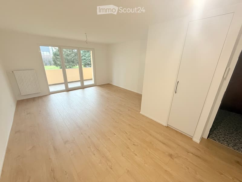 Empty room with light wooden flooring, white walls, large sliding glass doors leading to a balcony, and a radiator on the left wall.