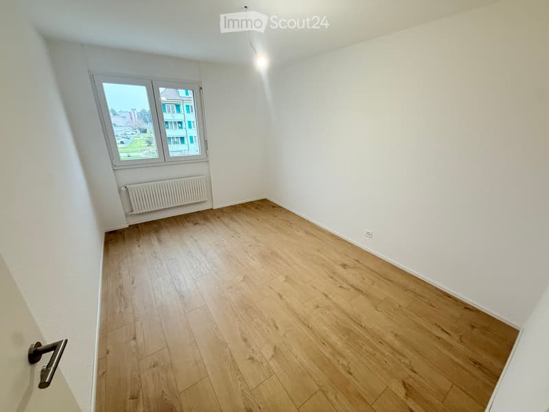 Empty room with light wooden flooring, white walls, and a window with a view of the outside. A radiator is visible below the window, and a door handle is seen on the left side.