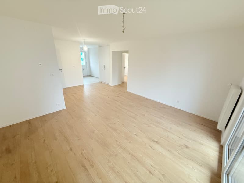 Empty room with light wooden flooring and white walls, featuring a doorway leading to another room and a window letting in natural light.