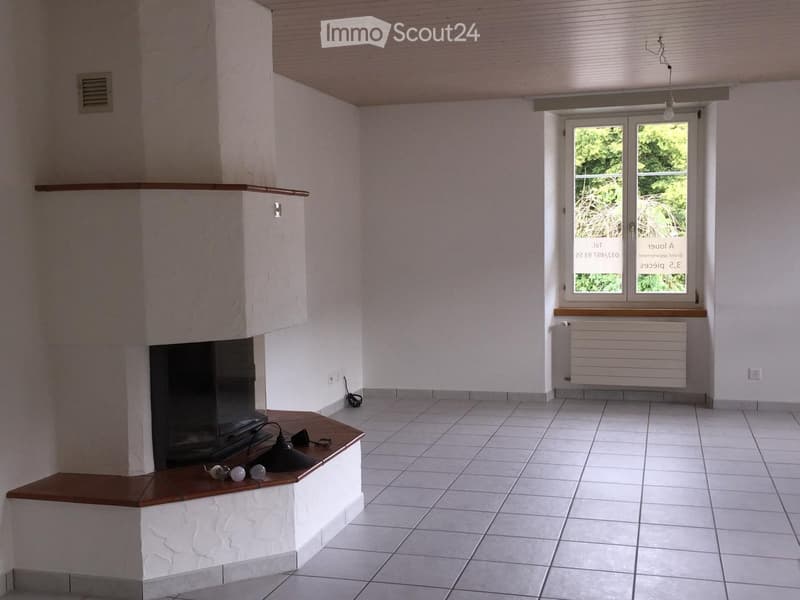 Empty room with white tiled floor, white walls, fireplace with wooden mantel, window with blinds, radiator, and visible electrical outlet.