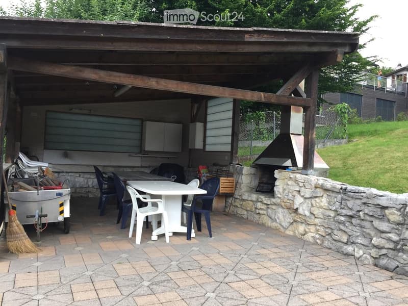 Wooden pergola-covered outdoor area with a stone wall, a white table with chairs, a barbecue, and a storage cart with firewood.