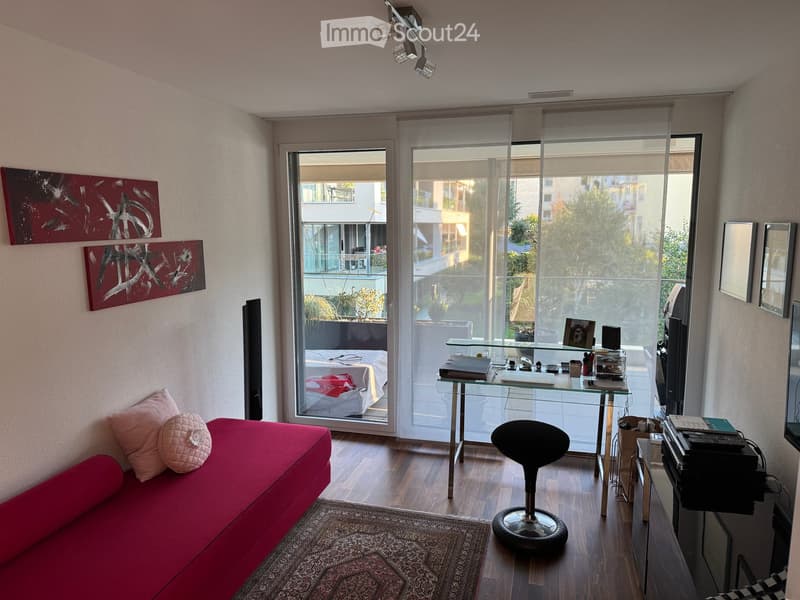 Modern living room with a red sofa, glass table, stool, laptop, and artwork on the wall. Large windows offer a view of a balcony and outdoor area.