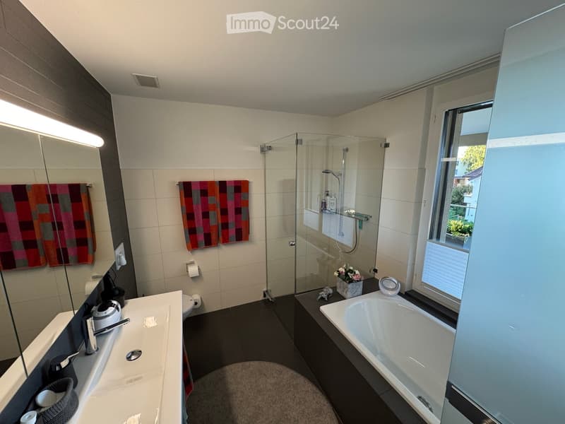 Modern bathroom with a bathtub, sink, mirror, and glass-enclosed shower. Features decorative towels, a flower vase, and a view through a window.