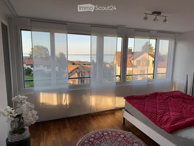 Bright bedroom with wooden flooring, large windows with sheer curtains, a bed with red bedding, a vase with white orchids, and a round patterned rug.