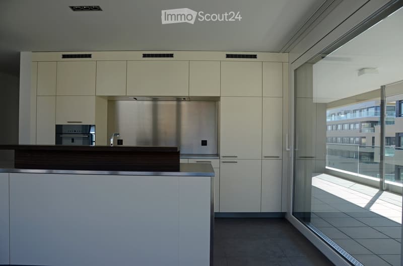 Modern kitchen with white cabinets, stainless steel appliances, and a dark wooden countertop. Large windows provide natural light and a view of the balcony.