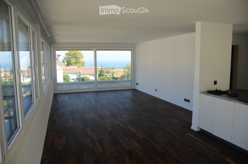 Empty room with dark wooden flooring, white walls, large windows, and a sliding glass door leading to a balcony with a scenic view.