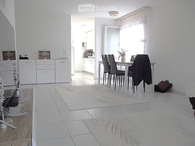 Bright, modern dining area with a white dining table and six chairs, a small kitchen area with white cabinets and appliances, and decorative elements including a vase with flowers and a suitcase on the floor.