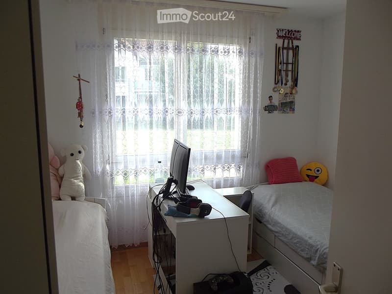 Cozy bedroom with a single bed, desk, and window with sheer curtains. The bed has pillows and stuffed toys, and the desk holds a computer setup with headphones.