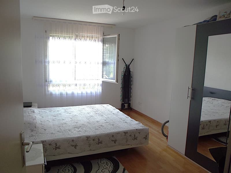Bedroom with a double bed, nightstand, mirror, and ironing board. Light-colored walls, wooden flooring, and a window with curtains.