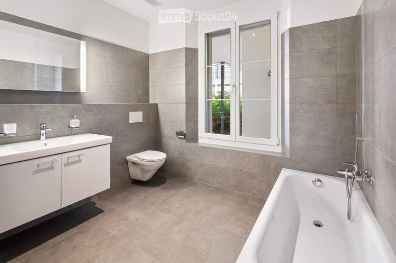 Modern bathroom with a bathtub, toilet, sink, and mirrored cabinet. Features grey tiles, a large window, and sleek fixtures.