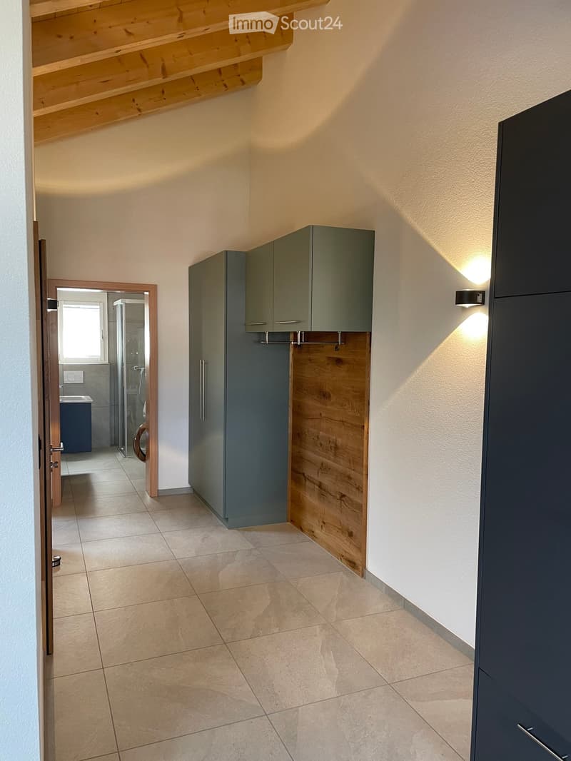 A modern hallway with tiled flooring, light-colored walls, and built-in grey cabinets with wooden accents. A small, open doorway leads to a bathroom with a visible sink and shower area.