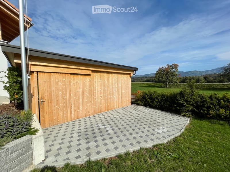 A modern wooden garage with a grey tiled driveway, surrounded by greenery and a scenic mountain view.