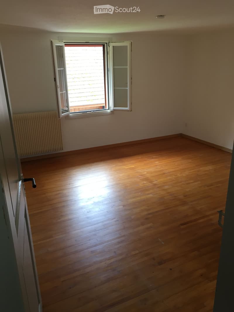 Empty room with wooden flooring, white walls, and a large window with blinds. The room has a radiator on one wall and a door with a handle visible on the left side.