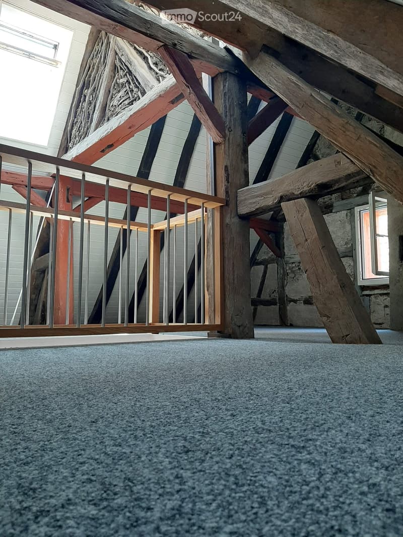 An attic space with exposed wooden beams, metal railing, and a carpeted floor. The room features large windows and a stone wall, with no furniture or furnishings present.