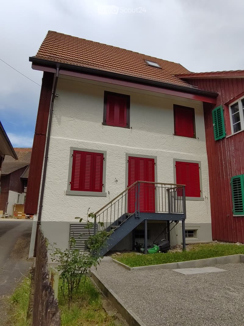 Two-story house with white walls, red shutters, and a small balcony accessed by stairs. A lawn and gravel driveway are visible in front.