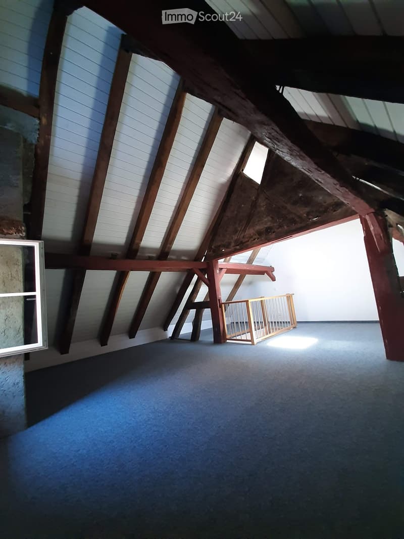 An empty attic space with exposed wooden beams, white walls, and a small window letting in natural light. The floor is carpeted, and there is a small wooden playpen in the corner.