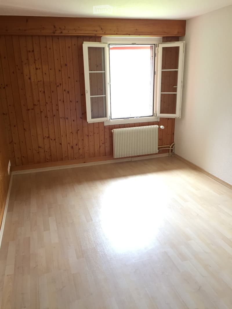 Empty room with wooden walls and flooring, featuring a large window with white frames and a radiator below it.