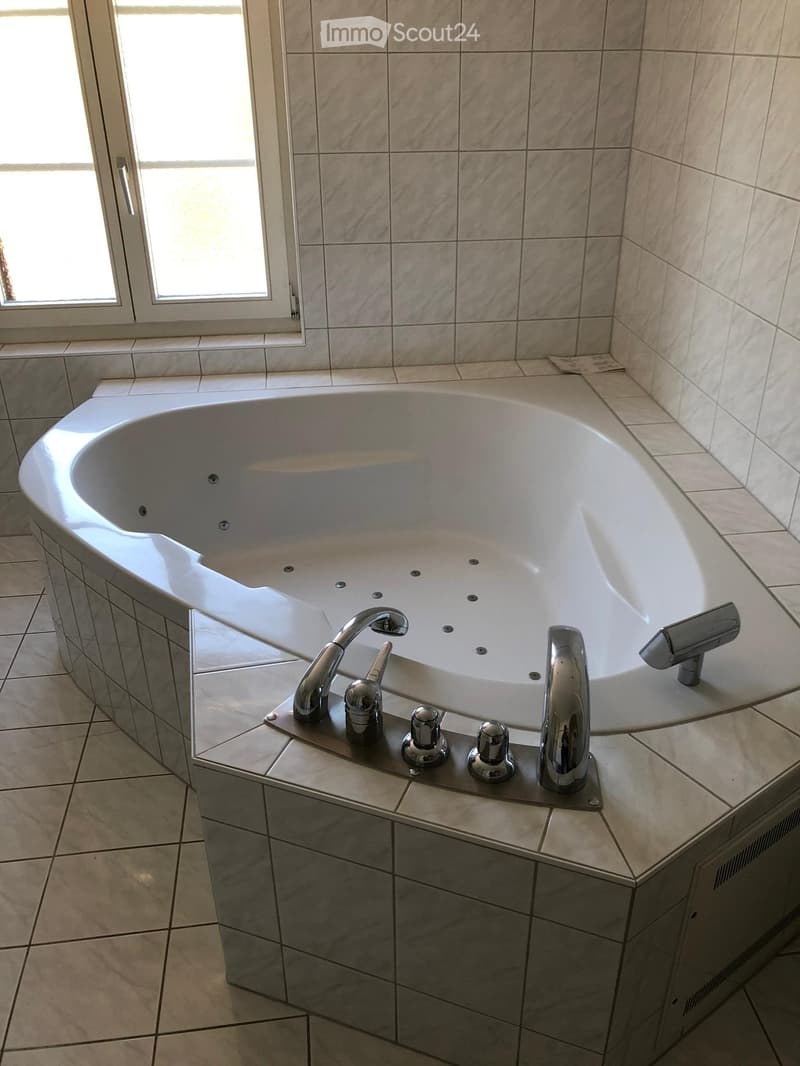 Modern bathroom with a large, oval bathtub, multiple chrome faucets, and tiled walls and floor. A window provides natural light.