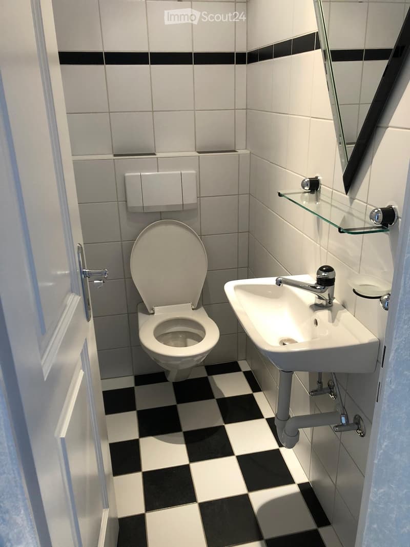 A compact bathroom with white tiles, black accents, a toilet, a sink with a faucet, a mirror, and a glass shelf.