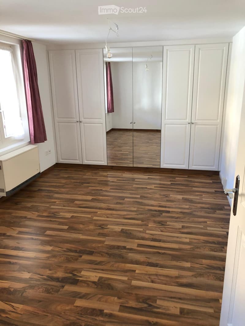 Empty room with wooden flooring, white walls, large mirrored closet, and window with curtains.