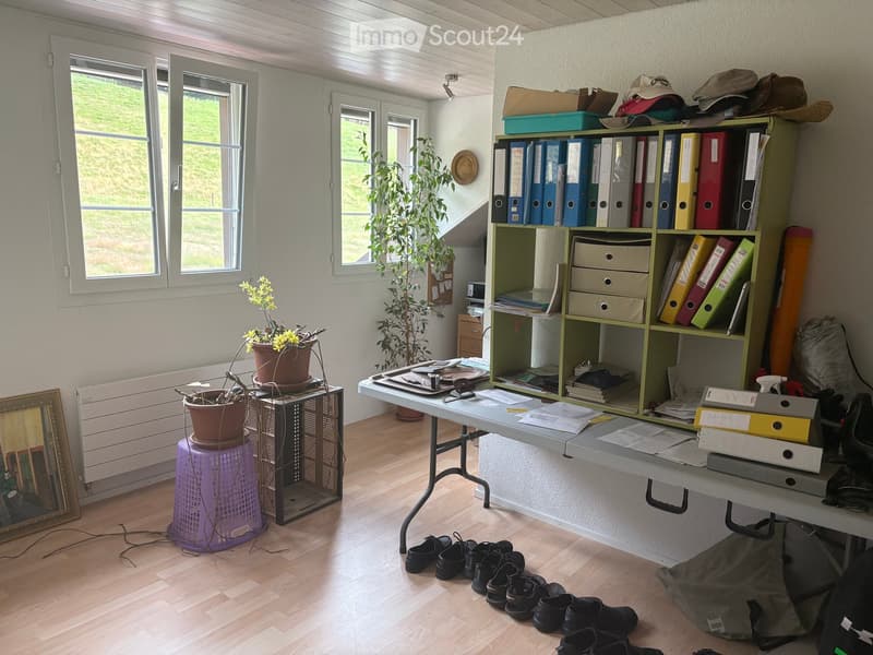 A well-lit room with wooden flooring, large windows, a table with office supplies, a shelf with binders and books, potted plants, a framed picture, and a purple stool with a potted plant on top.