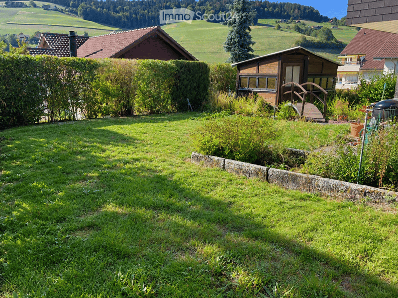 Lush green garden with a stone border, small wooden structure, and surrounding houses in a rural landscape.