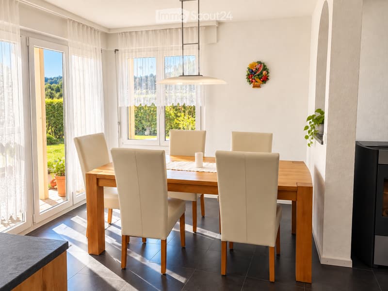 Bright dining room with a wooden dining table surrounded by white leather chairs, a candle in the center, large windows with sheer curtains, and a decorative wall piece.