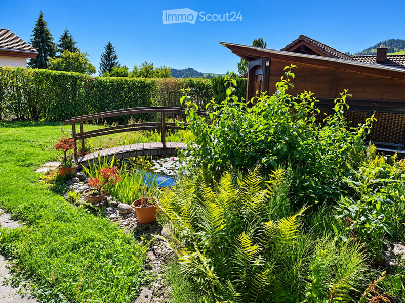A well-maintained garden with a wooden bridge over a small pond, surrounded by lush greenery, ferns, and potted plants. The area features a wooden house in the background and a clear blue sky overhead.