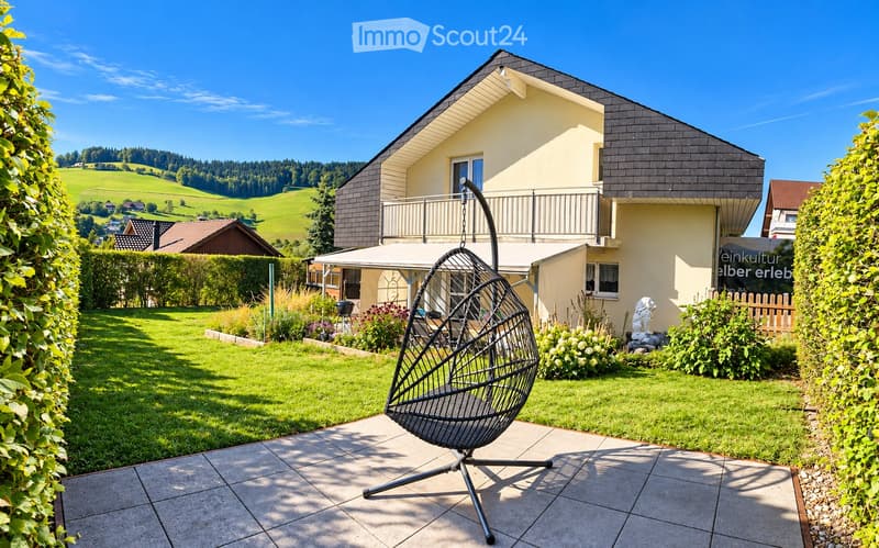 Well-maintained garden with a modern house in the background, featuring a wicker hanging chair, lush greenery, and a scenic countryside view.