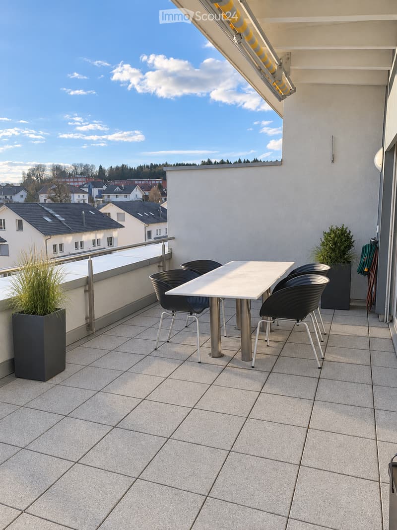 Balcony with tiled floor, white table, four black chairs, and two potted plants, overlooking a residential area with houses and trees.