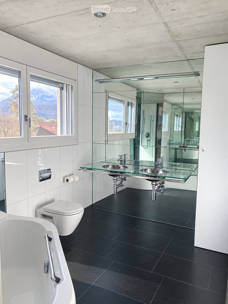 Modern bathroom with a bathtub, toilet, double sink with glass countertop, large mirror, and scenic mountain view through large windows.