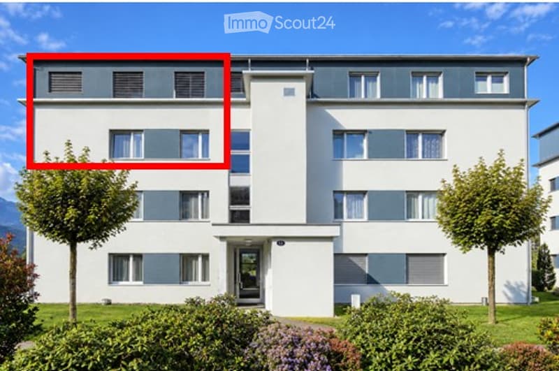 Modern multi-story apartment building with white facade, grey accents, and multiple windows. Features a central entrance and landscaped surroundings with trees and shrubs.