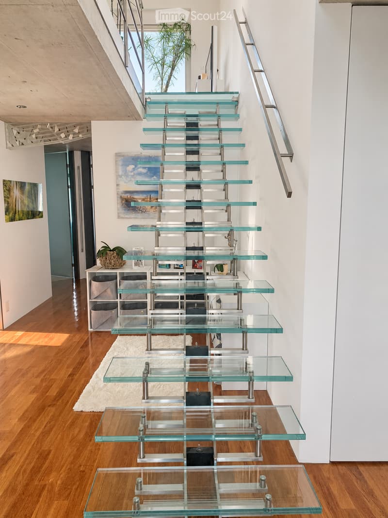 Modern hallway with a sleek glass staircase featuring metal railings, wooden flooring, and contemporary decor including a small shelf unit and artwork.