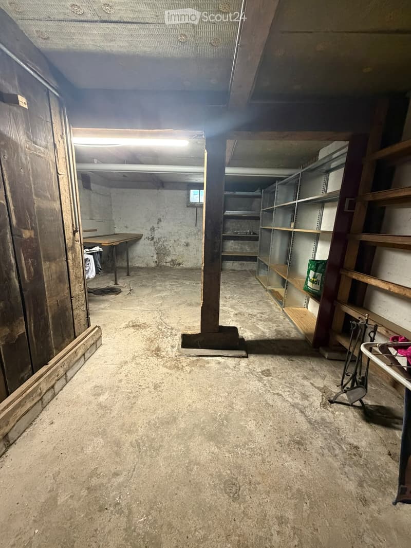 Empty basement space with concrete floor, exposed wooden beams, metal shelving units, and a wooden table with a few items on it.