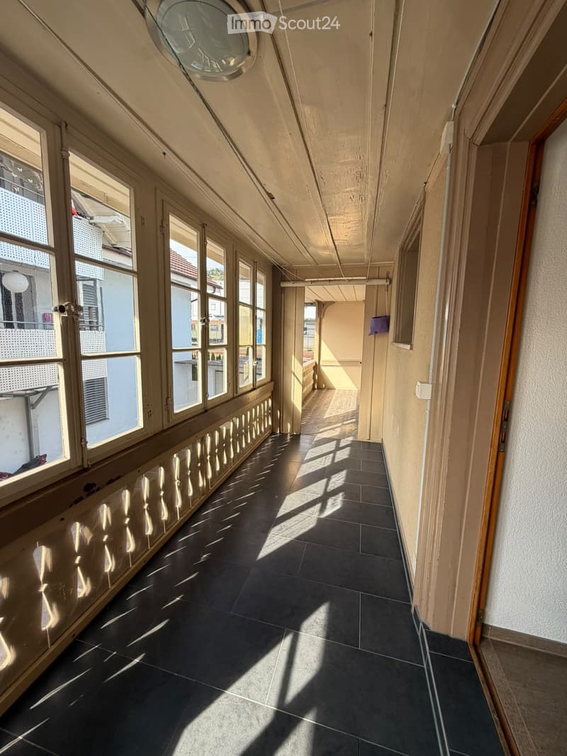 A long, narrow hallway with dark tiled flooring and large windows on one side, allowing natural light to illuminate the space. The walls are painted in a light color, and the ceiling features exposed wooden beams and a skylight.