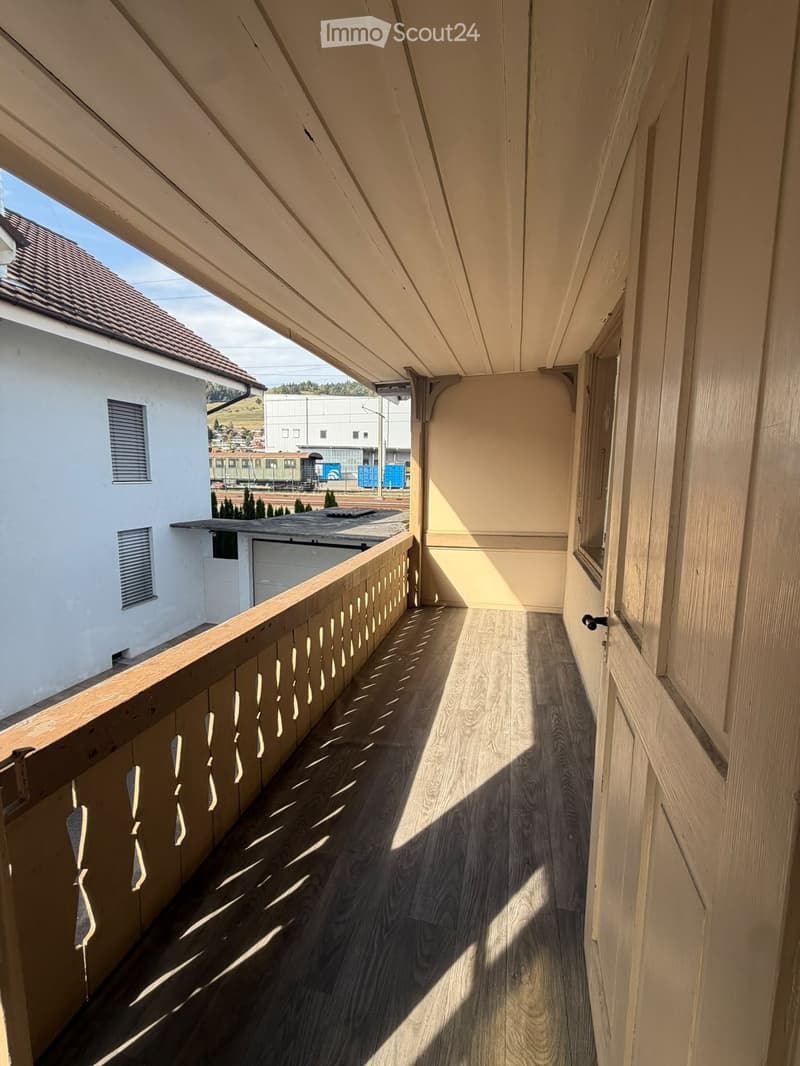Wooden balcony with a railing, wooden floor, and a view of a neighboring building and landscape.