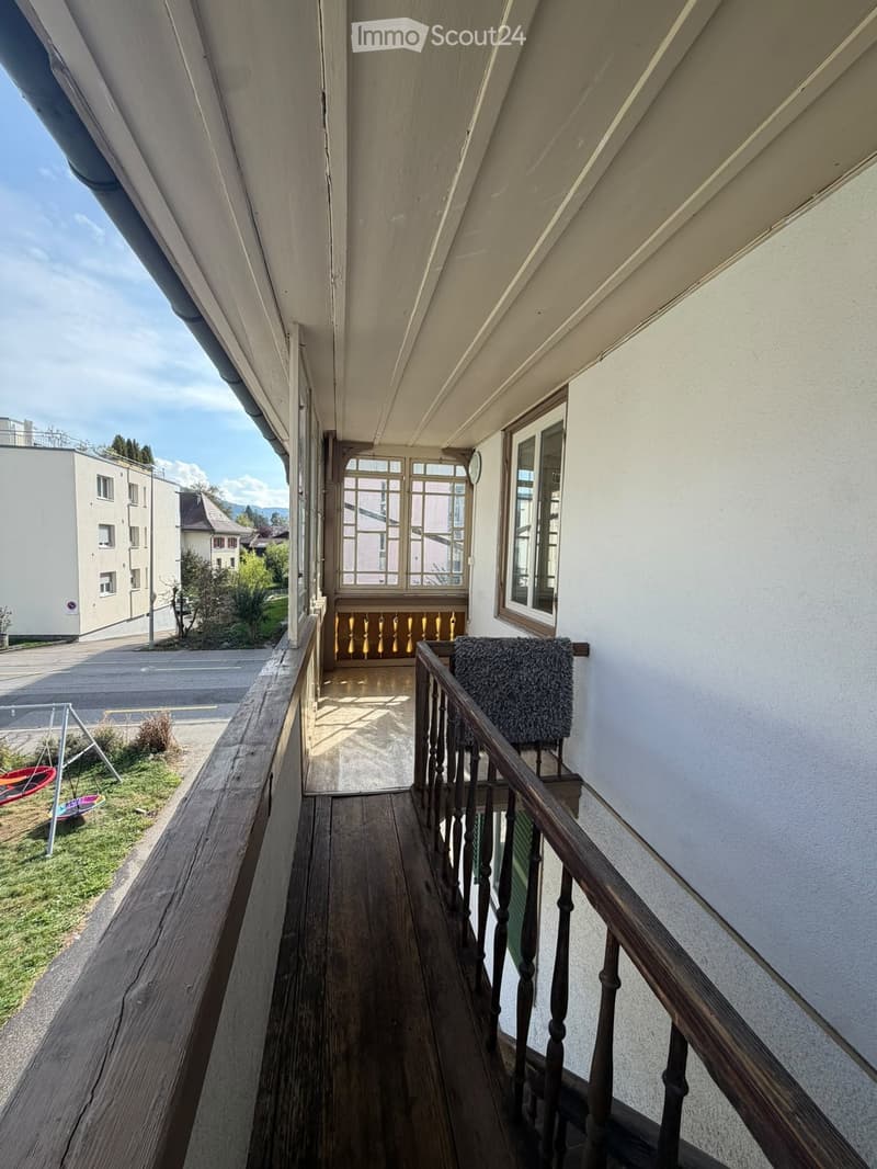 Wooden balcony with railing, leading to a doorway with glass panels and decorative balusters.