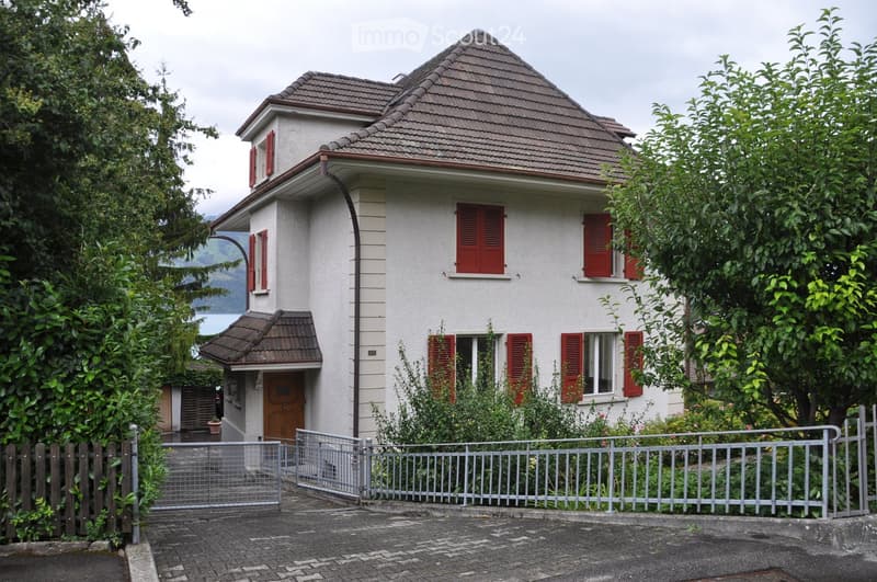 Two-story house with white exterior, red shutters, and a brown tiled roof. The house features a gated entrance, a small porch, and is surrounded by greenery and trees.