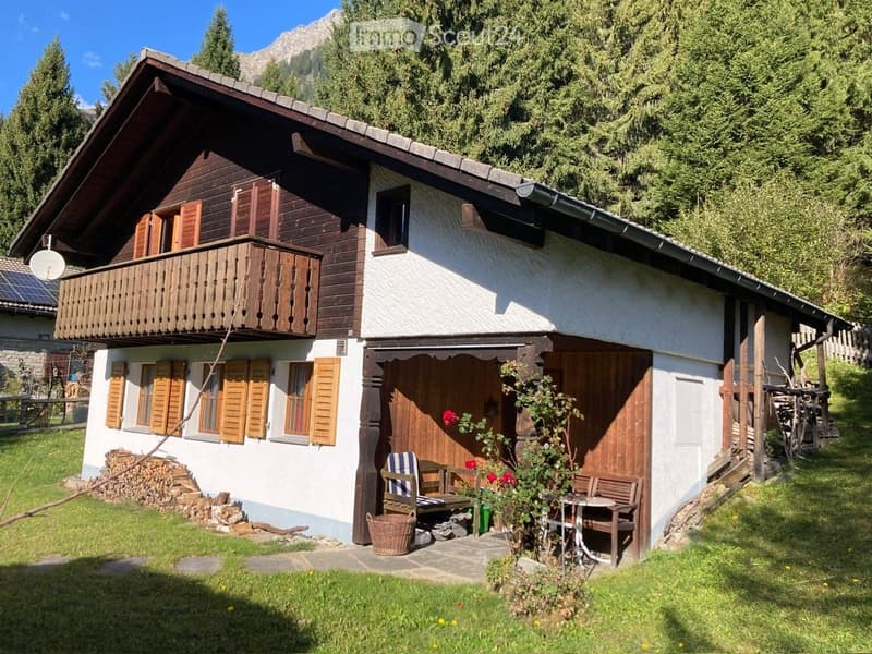 A two-story wooden house with white walls, wooden shutters, and a balcony on the upper floor. The house features a small porch with wooden benches, a table, and a wicker basket, surrounded by a grassy yard with a stack of firewood and a view of mountains
