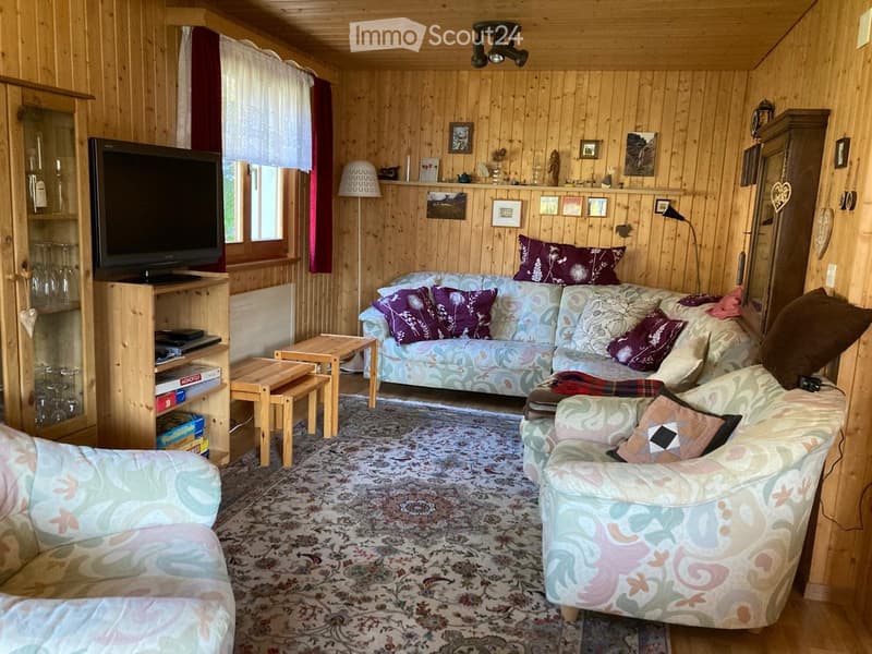 Cozy living room with wooden walls, a patterned rug, a floral-patterned sofa, armchairs, a coffee table, a television, and a wooden cabinet with glass doors.