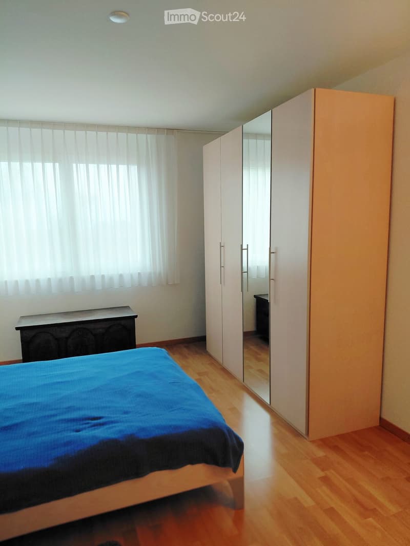 Spacious bedroom with wooden flooring, a large bed with blue bedding, a wardrobe with mirrored doors, and a small table near the window.