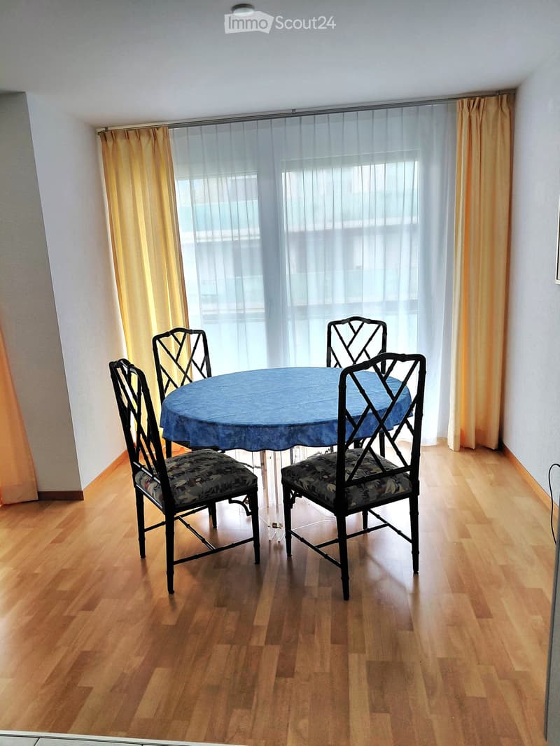Dining area with a round table covered in a blue tablecloth and four matching chairs with floral upholstery, set against wooden flooring and large windows with sheer curtains.