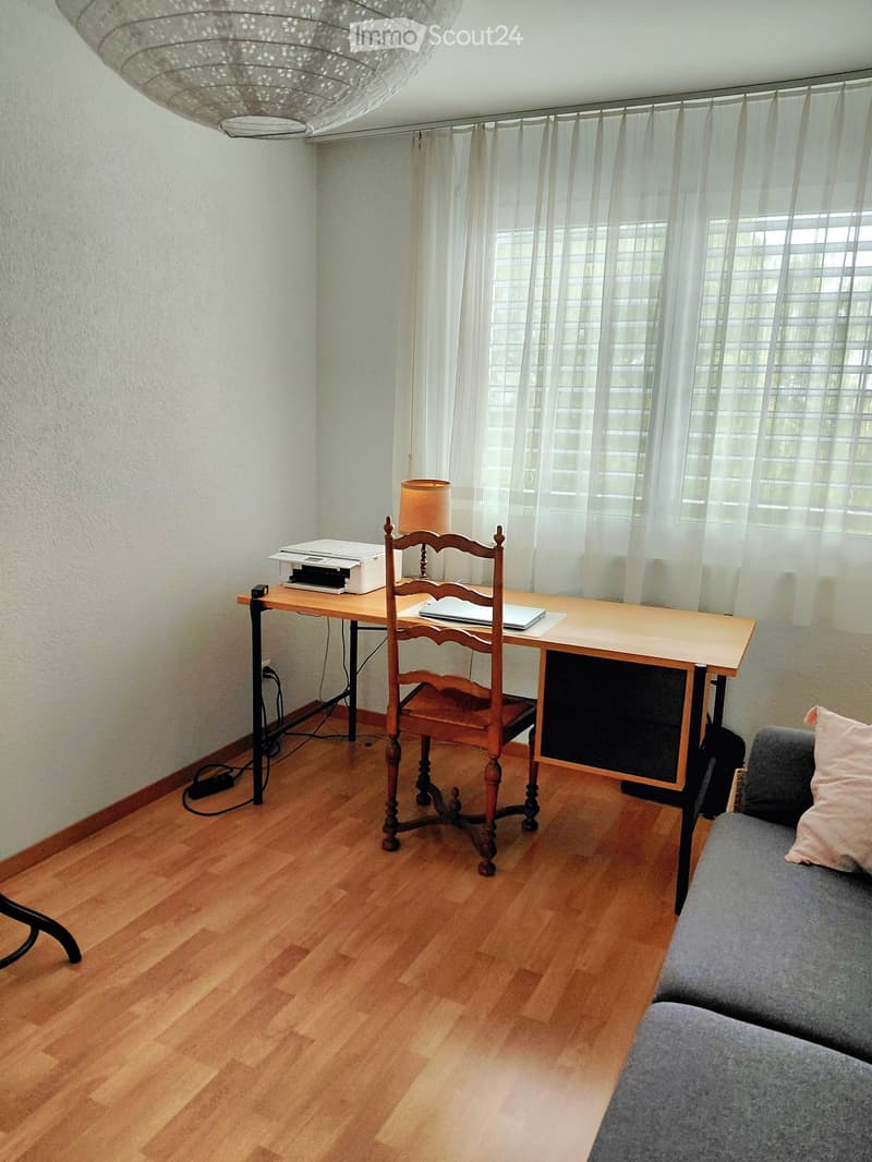A well-lit room with wooden flooring, a grey sofa with a pink pillow, a wooden desk with a chair, a printer, a laptop, and a lamp on the desk. Large windows with white curtains allow natural light to fill the space.
