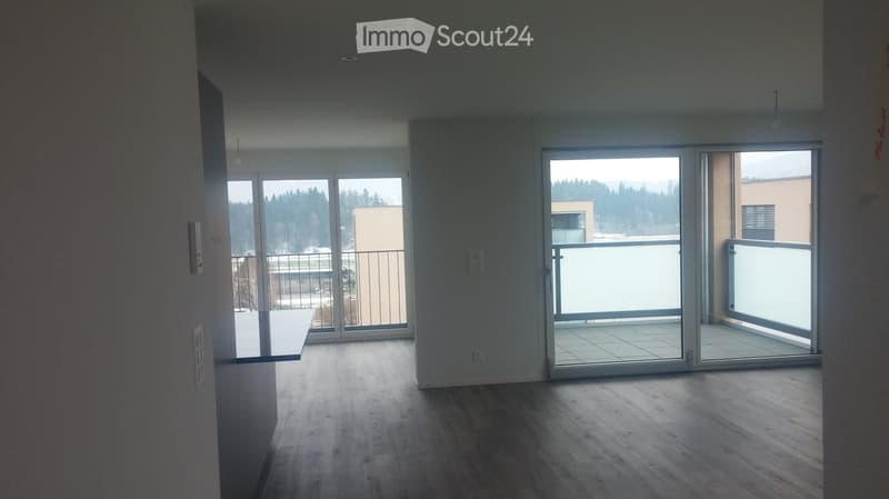 Empty room with wooden flooring, large sliding glass doors leading to a balcony, and a small table near the left wall.