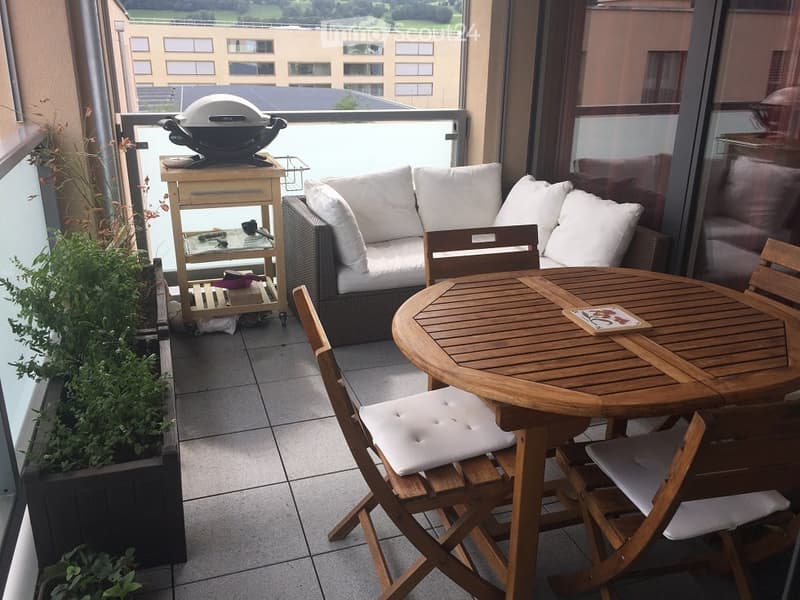Balcony with a round wooden table, four folding chairs, a wicker sofa, a small cart with a grill, and potted plants.