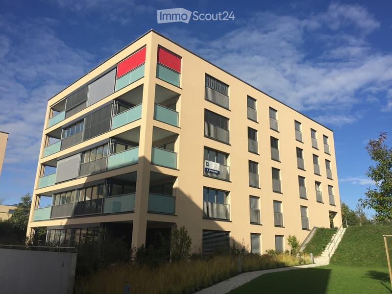 Modern multi-story residential building with light beige facade, large windows, and balconies. Features a raised ground floor and a sign reading "A VENDRE" (For Sale).
