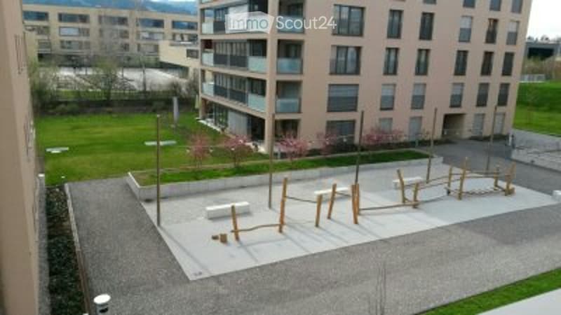 Outdoor playground area with wooden play structures, benches, and open space surrounded by modern apartment buildings.
